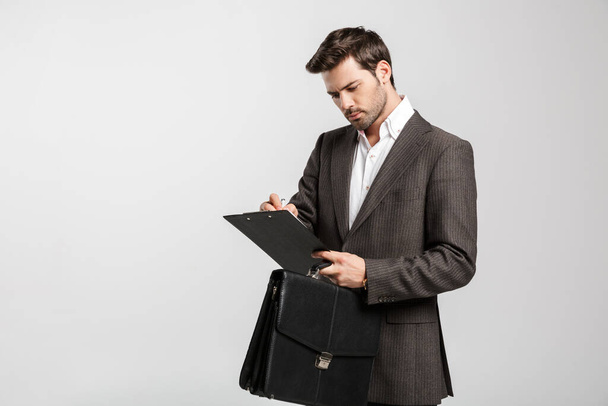 Photo of a handsome serious young businessman isolated over white wall background holding bag and clipboard. - Foto, imagen
