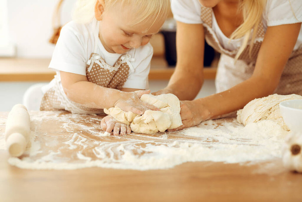 Little girl and her blonde mom in beige aprons playing and laughing while kneading the dough in kitchen. Homemade pastry for bread, pizza or bake cookies. Family fun and cooking concept - Zdjęcie, obraz