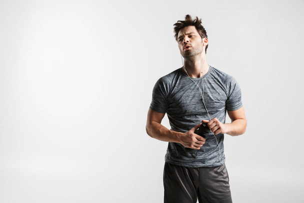 Image of young tired man using earphones and holding water bottle while working out isolated over white background - Fotó, kép