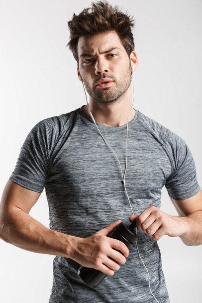 Image of young tired man using earphones and holding water bottle while working out isolated over white background - Foto, afbeelding