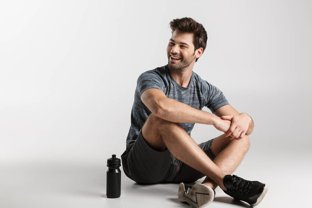 Smiling young sportsman sitting isolated over gray background, resting after workout, looking away - Foto, Bild