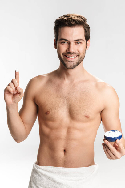 Zdjęcie radosnego półnagiego mężczyzny w ręczniku kąpielowym uśmiechającego się i pokazującego krem do twarzy odizolowany na białym tle - Zdjęcie, obraz