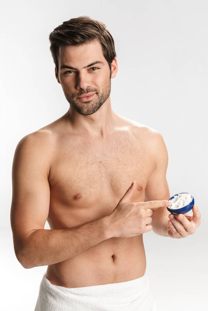 Photo of handsome half-naked man in bath towel applying facial cream isolated over white background - Foto, afbeelding