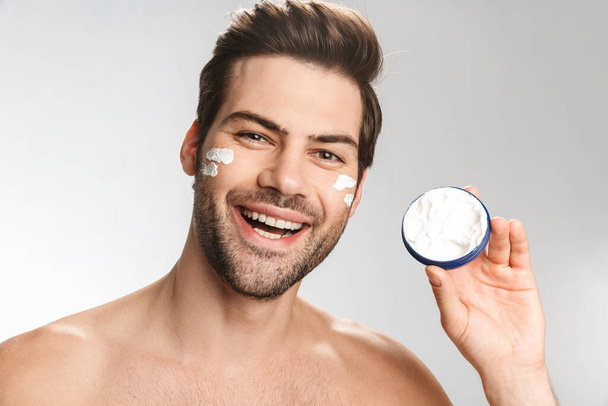 Photo of joyful half-naked man smiling and showing facial cream isolated over white background - Φωτογραφία, εικόνα