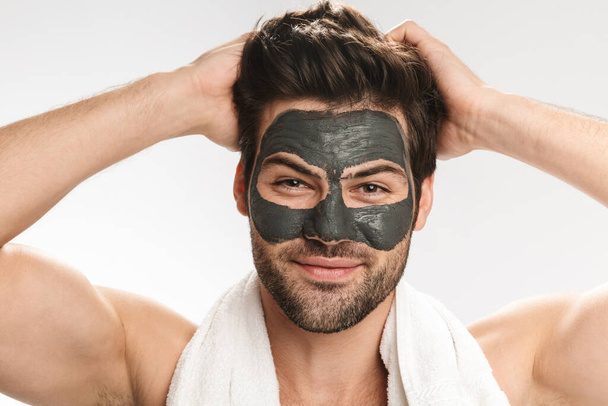 Photo of happy half-naked man with cosmetic clay mask on his face and towel smiling at camera isolated over white background - Φωτογραφία, εικόνα