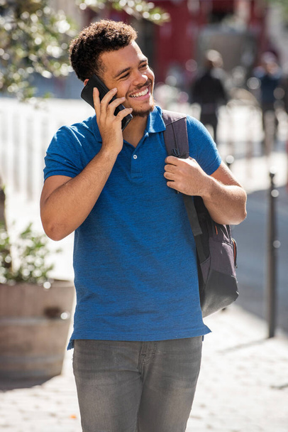Portret van een lachende jonge Afro-Amerikaanse man die loopt en praat met mobiele telefoon en tas - Foto, afbeelding