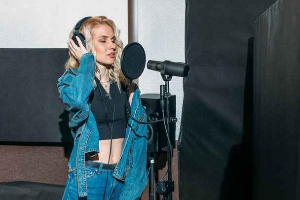young beautiful girl, blonde, singer, makes a recording in a recording studio, she stands next to the microphone on a dark background, she has a beautiful smile and emotions. a sound engineer sits at a computer and monitors the process - Foto, afbeelding