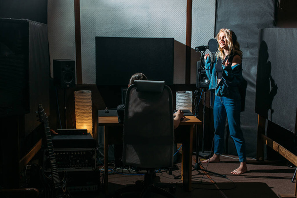 young beautiful girl, blonde, singer, makes a recording in a recording studio, she stands next to the microphone on a dark background, she has a beautiful smile and emotions. a sound engineer sits at a computer and monitors the process - Φωτογραφία, εικόνα