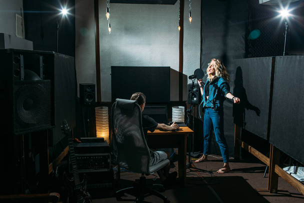 young beautiful girl, blonde, singer, makes a recording in a recording studio, she stands next to the microphone on a dark background, she has a beautiful smile and emotions. a sound engineer sits at a computer and monitors the process - Fotó, kép