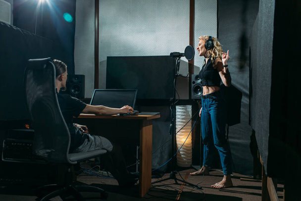 young beautiful girl, blonde, singer, makes a recording in a recording studio, she stands next to the microphone on a dark background, she has a beautiful smile and emotions. a sound engineer sits at a computer and monitors the process - Photo, Image