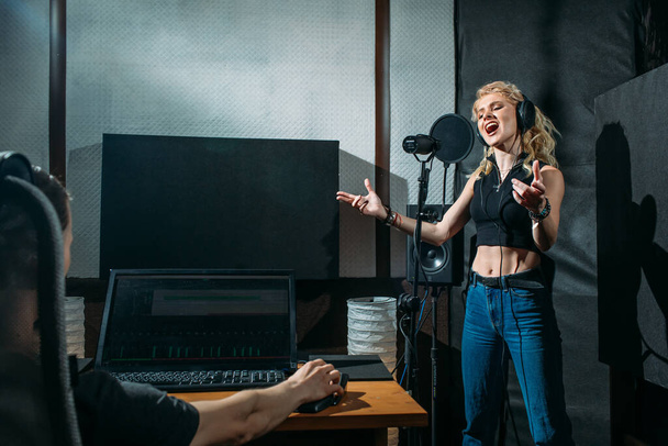 young beautiful girl, blonde, singer, makes a recording in a recording studio, she stands next to the microphone on a dark background, she has a beautiful smile and emotions. a sound engineer sits at a computer and monitors the process - Фото, зображення