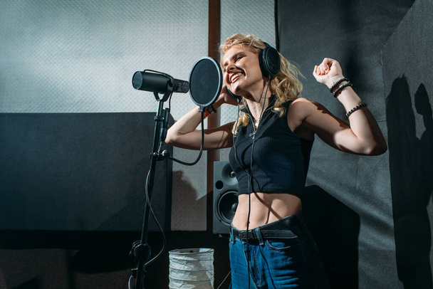 young beautiful girl, blonde, singer, makes a recording in a recording studio, she stands next to the microphone on a dark background, she has a beautiful smile and emotions. a sound engineer sits at a computer and monitors the process - Фото, изображение