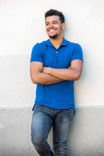 Retrato de un joven mestizo sonriente apoyado en una pared blanca con los brazos cruzados
 - Foto, Imagen