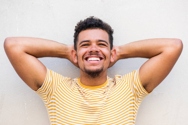 Primer plano retrato de joven afroamericano guapo riendo con las manos detrás de la cabeza contra la pared blanca
 - Foto, imagen