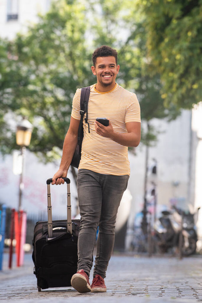 Full body portrait of happy young man travel with cellphone and suitcase on street - Foto, Bild