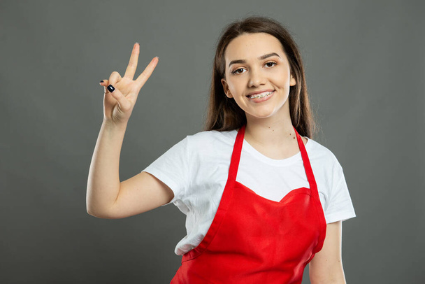 Portrét mladé atraktivní zaměstnankyně supermarketu ukazující mírové gesto na šedém pozadí - Fotografie, Obrázek