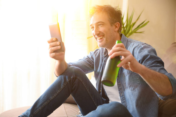 Чоловік посміхається п'є пиво з пляшки і перекушує сидячи на дивані у вітальні вдома
 - Фото, зображення