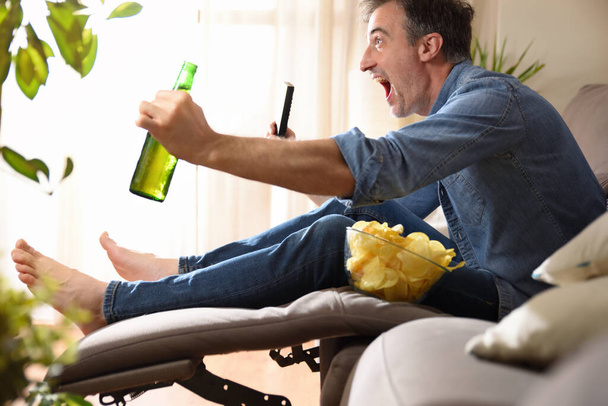 Man watching sports on television sitting comfortably on a sofa having a snack with beer and a remote control in his hands. - Fotoğraf, Görsel
