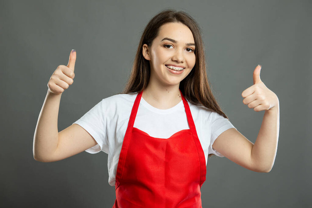Muotokuva nuori nainen supermarket työntekijä osoittaa kaksinkertainen peukalo ylös ele harmaalla pohjalla - Valokuva, kuva