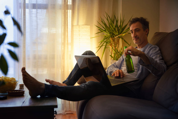 Relaxed man entertaining with a laptop at home having a beer sitting on a sofa - Фото, зображення