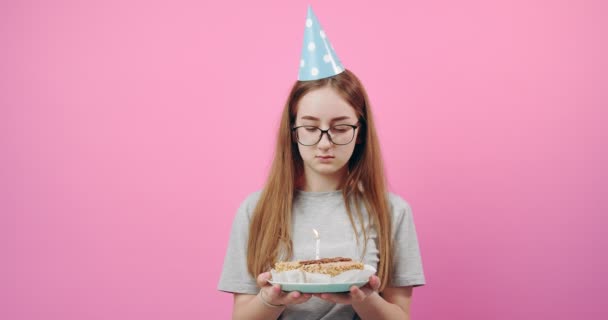 Upset girl in festive cap holding cake with candle in studio - Кадри, відео