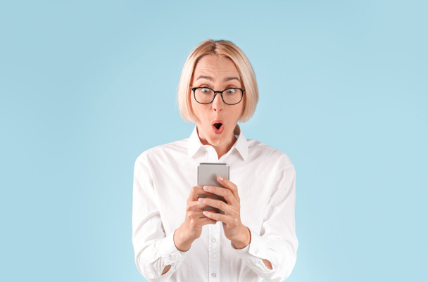 Sorprendida dama de negocios gritando OMG mientras mira el teléfono inteligente contra el fondo azul
 - Foto, imagen