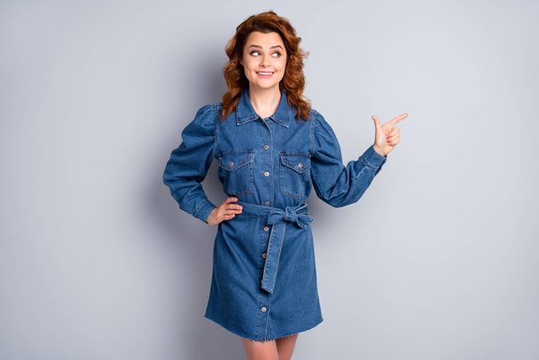 Photo of funny ginger curly lady directing finger side empty space showing novelty sale price product black friday wear casual blue denim short dress isolated grey color background - Foto, Imagem