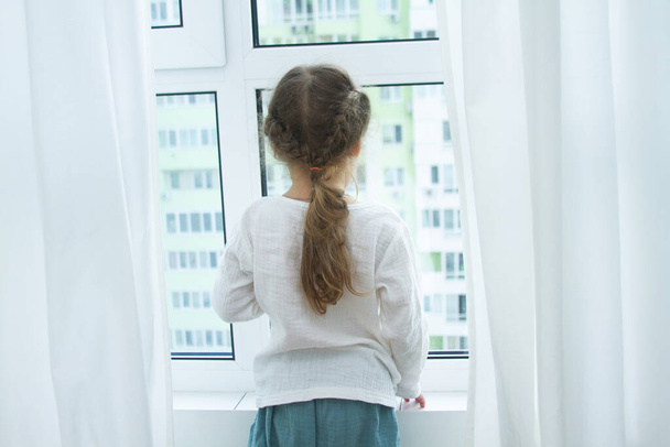 Menina bonito em um quarto brilhante, um mínimo de objetos na sala, a criança se alegra, sentado à janela com cortinas brancas
. - Foto, Imagem