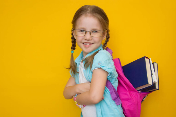 Back to school. Beautiful little girl in glasses and with pigtails with books in a school backpack smiling looking at the camera on a yellow background. Education concept. - Foto, imagen