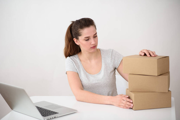 Девушка с хвостиком, серая футболка сидит за ноутбуком и смотрит на стопку картонных коробок. Белый фон. Сервис доставки онлайн
 - Фото, изображение