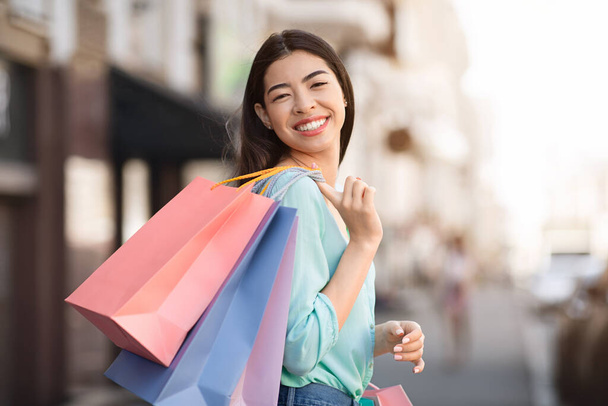 Gülümseyen Asyalı kadın dışarıda alışveriş torbaları taşıyor ve kameraya poz veriyor. - Fotoğraf, Görsel