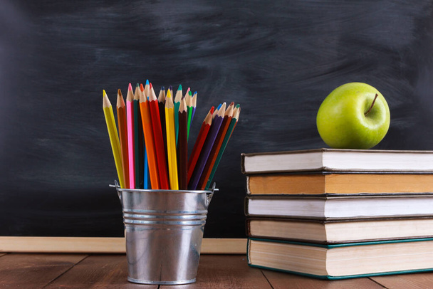 Colorful pencils on brown wooden desk. Green apple on stack of books. Classroom blackboard  in background. Education, back to school, autumn, September concept - Photo, image