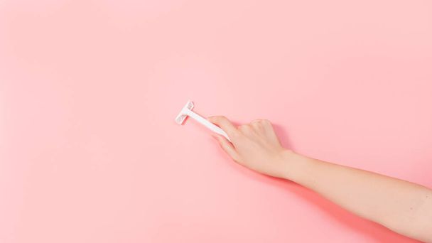 Female holding white razor against pink colored background. Removal hair. Minimalism. Top view. New disposable razor. Body hygiene, spa procedures banner - 写真・画像