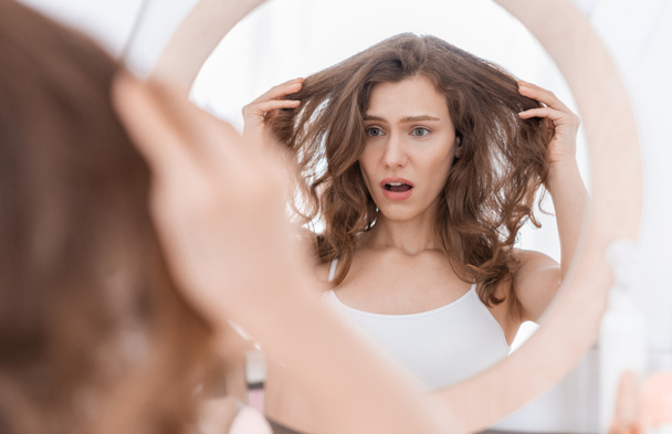 Schockierte junge Frau hält sich die Haare und blickt in den Spiegel - Foto, Bild