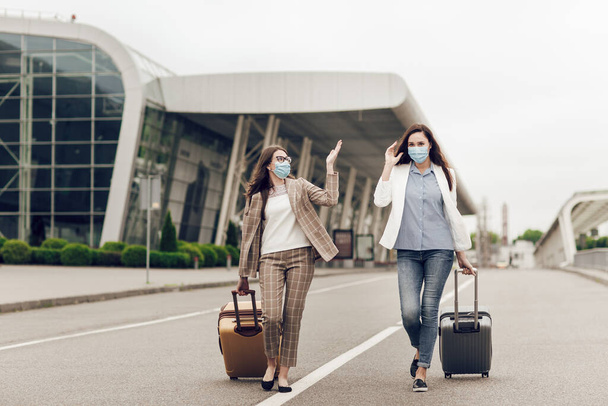 Deux femmes heureuses portant des masques de protection après une quarantaine de coronavirus avec des valises se rendent à l'aéroport. Jeunes femmes près de l'aéroport, vol d'ouverture, concept de voyage - Photo, image