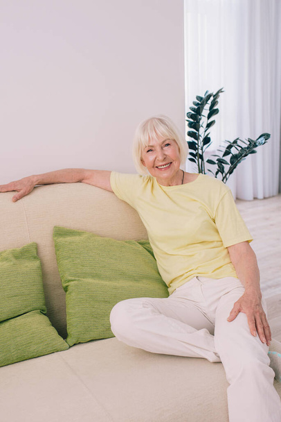 Yaşlı (yaşlı) neşeli beyaz kadın, beyaz saçlı, evdeki kanepede oturuyor. Yaş karşıtı, sağlıklı yaşam tarzı, pozitif düşünce konsepti. - Fotoğraf, Görsel
