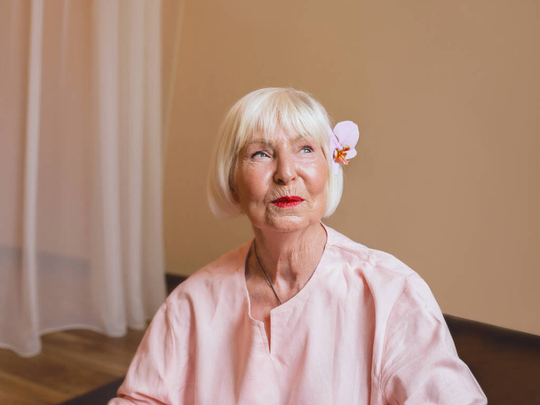 elderly (old) caucasian stylish woman with gray hair and pink phalaenopsis in her hair sitting at ashram with namaste hands. Anti age, healthy lifestyle, monk, thai massage concept - Photo, Image