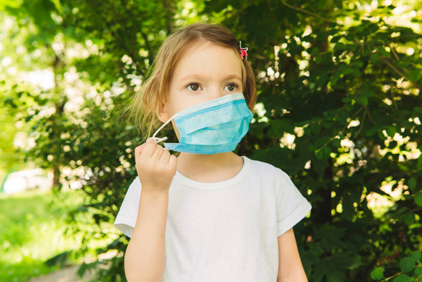 Little cute girl take off medical face mask in the outdoor, trees on background, close up. - Фото, изображение