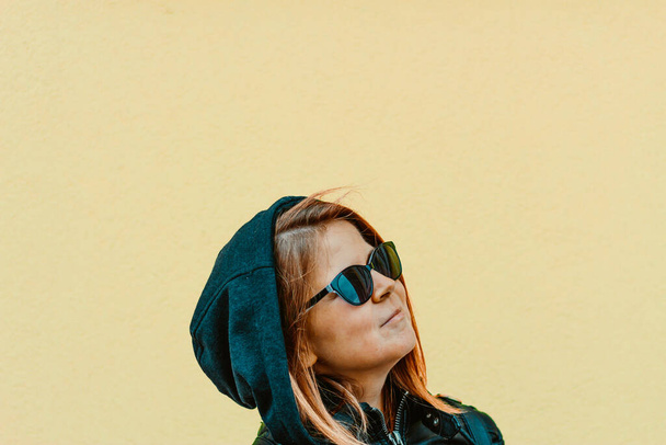 Молодая женщина в сером капюшоне и солнечных очках улыбается на ярко-желтом фоне Брюнетка в городской одежде и черных очках выглядит уверенно и вверх
 - Фото, изображение