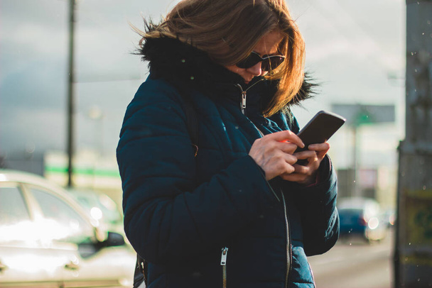 Молода жінка в густому одязі та сонцезахисних окулярах тримає свій телефон Гарненька леді з мобільним пристроєм, використовуючи його для смс, спілкування та соціалізації
 - Фото, зображення