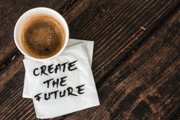 A full cup of coffee with a foam sitting on a white napkin with the message create the future handwritten on it placed on a wooden surface Concept image for inspirational messages - Photo, Image
