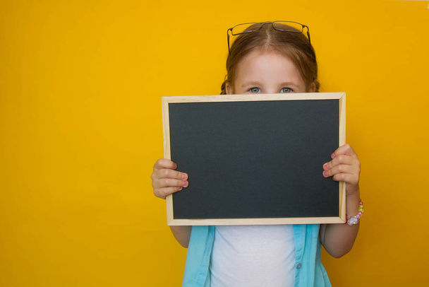 Portrait of a beautiful little girl peeking from behind a blackboard in her hands, smiling on a yellow background. Attractive positive baby. back to school. - 写真・画像