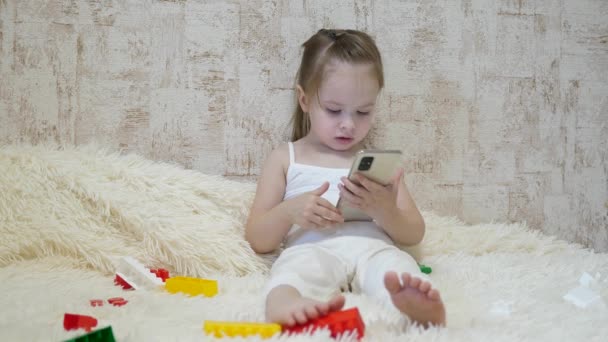 Dziecko bawi się przez telefon siedząc na kanapie z projektantem. Zabawka w smartfonie. Życie jako edukacja przedszkolna na odległość. Nowoczesne dziecko - Materiał filmowy, wideo