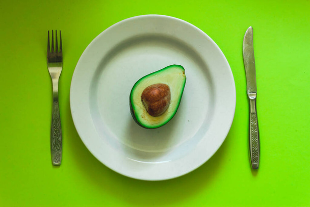 Half of green and ripe avocado with seed sitting on a white large plate with knife and fork on aside Minimalistic dish from a vegan diet full of oils and nutrients - Foto, Imagen