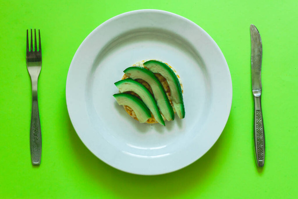 Krajíc zeleného a zralého avokáda na kusu kulatého rýžového chleba sedícího na velkém bílém talíři s vidličkou a nožem vedle něj Koncepční obraz pro zdravé stravování a vegetariánskou stravu - Fotografie, Obrázek