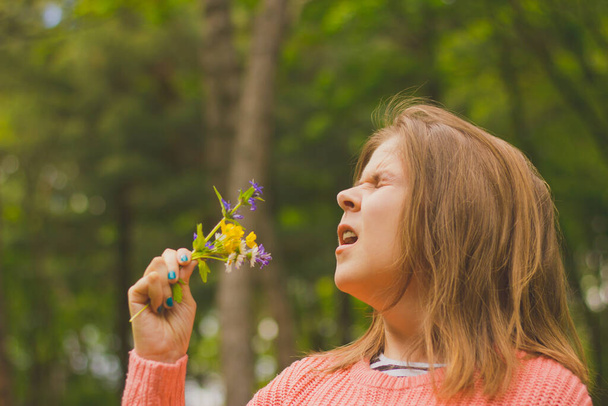 Mujer joven con cabello castaño usando una blusa rosa que tiene una reacción alérgica al polen Chica hipersensible estornudando mientras sostiene un ramo de flores
 - Foto, Imagen