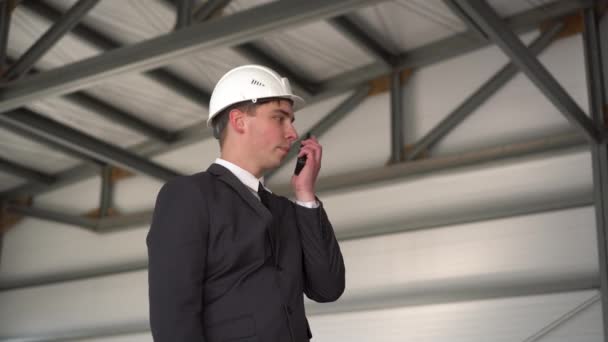 Ein junger Mann mit Helm spricht auf einer Baustelle auf einem Walkie-Talkie. Der Chef im Anzug schaut sich um. - Filmmaterial, Video