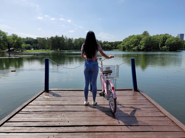 La joven morena con bicicleta rosa se para en un puente de madera y medita en el bosque a orillas del lago. Día soleado de verano. Fin de semana relajante
. - Foto, imagen