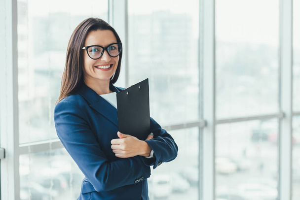 Confident young businesswoman standing with folder in hands, looking at the camera, ready to hard work day. - Foto, Imagen