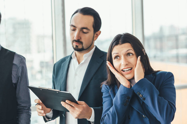 Shocked businesswoman, keeping hands on cheeks, looking at the camera, expressing despair and surprise while serious director is reading a report. - Foto, imagen